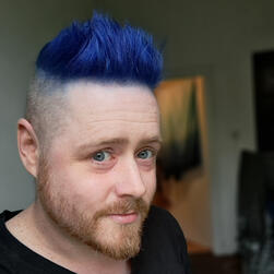 a selfie. i am white with blue hair spiked up to a point with shaved sides and a mid length orange beard. i'm on a boat at sea at sunset and am smiling at the camera