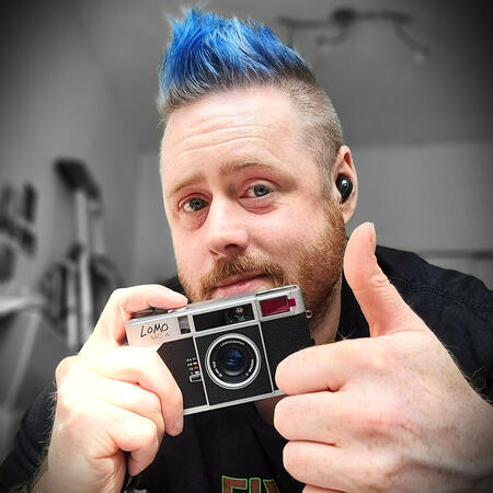 a selfie of a freckled white person with an orange beard flecked with white, and a blue faux hawk, shaved at the sides. i am smiling and holding a lomo mc-a black and silver point and shoot camera while also holding my other thumb up.