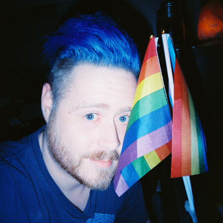 a selfie of a freckled white person with an orange beard flecked with white, and a blue faux hawk, shaved at the sides. i am smiling next to a pair of small pride flags.