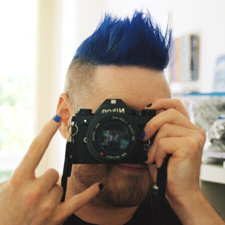 a selfie of a freckled white person with an orange beard flecked with white, and a blue faux hawk, shaved at the sides. i am smiling next to a pair of small pride flags.