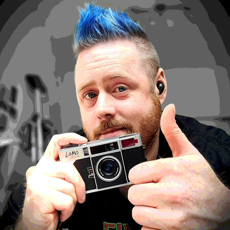 a selfie of a freckled white person with an orange beard flecked with white, and a blue faux hawk, shaved at the sides. i am smiling and holding a lomo mc-a black and silver point and shoot camera while also holding my other thumb up.
